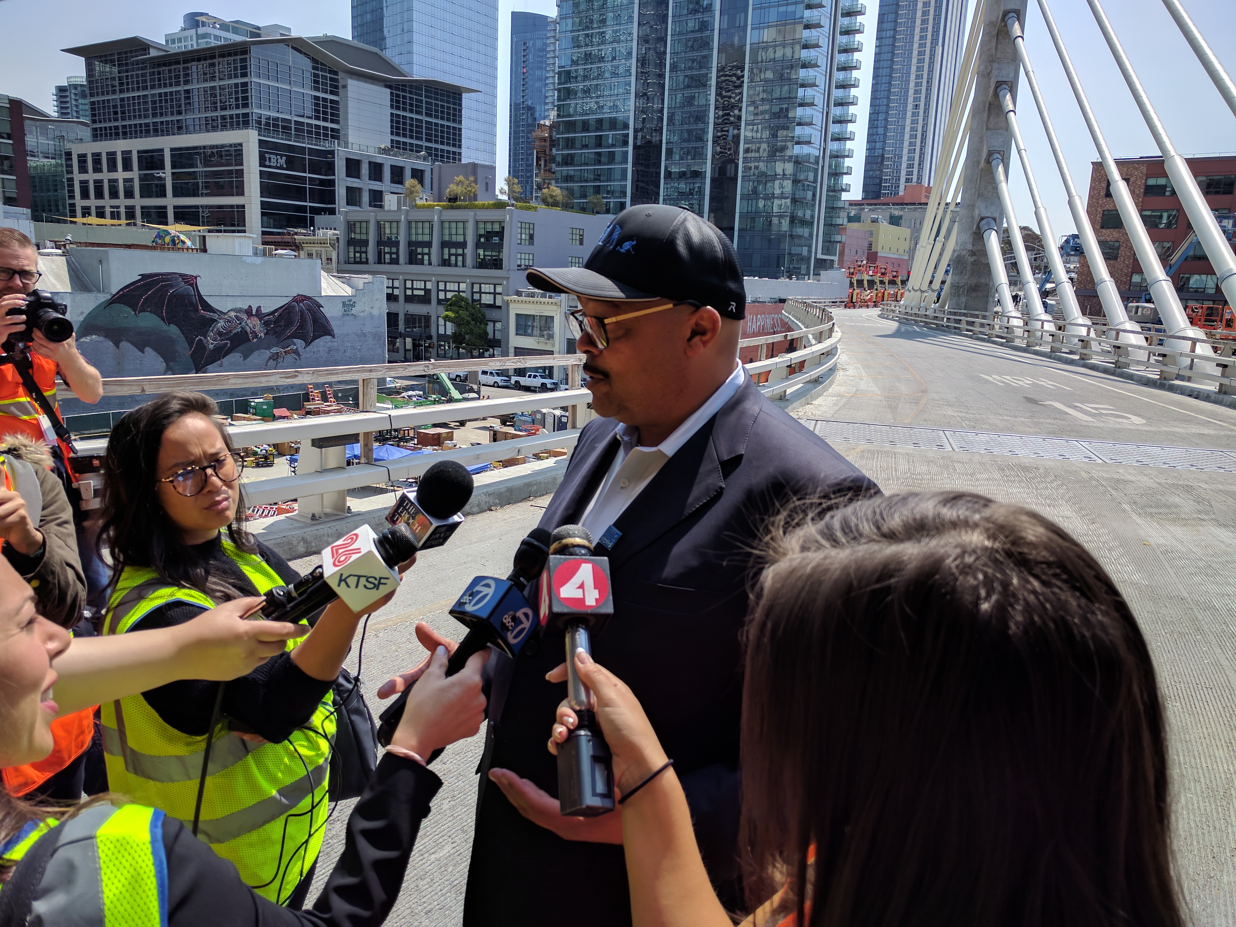 Nuru addressing the press with the bus bridge to the Bay Bridge behind him